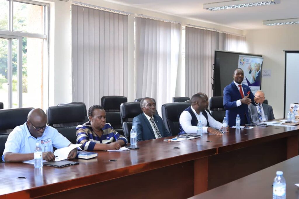 Six people sit at a conference table with notebooks and water bottles, while one man stands and speaks about the Transforming Education Program; a presentation screen in the background highlights its impact in Uganda.