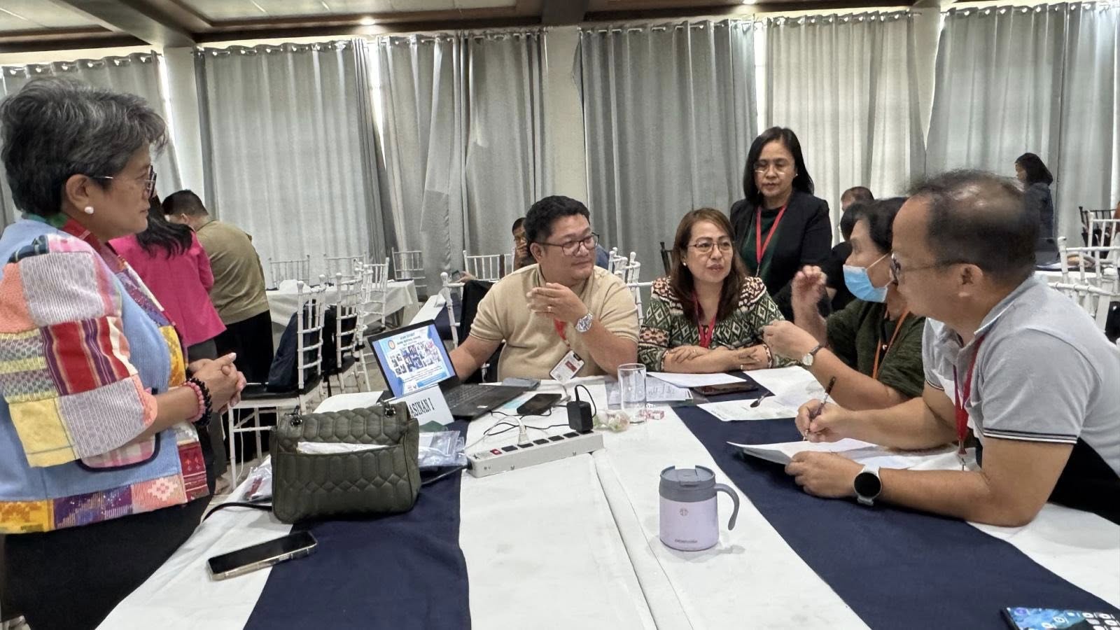 A group of people sit and stand around a table in discussion at an indoor event, focusing on holistic leadership development, with papers, mugs, and bags on the table.