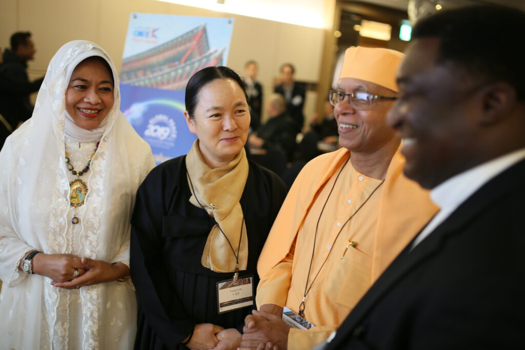 Four people wearing diverse traditional and religious attire stand together indoors, smiling and conversing at an event celebrating Interfaith Harmony. A poster and other attendees are visible in the background.