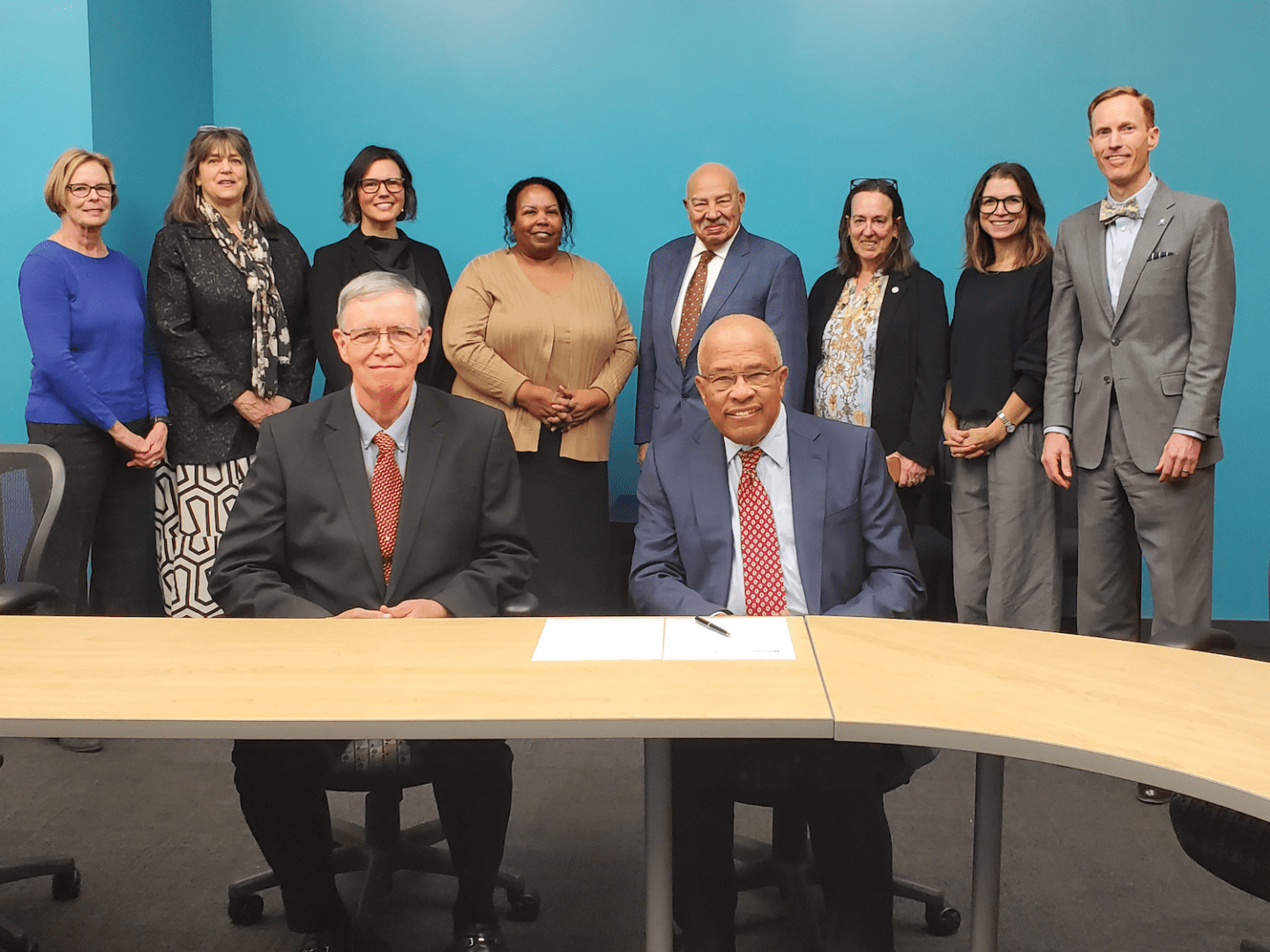 Two men sit at a table with papers in front of them, while nine people stand behind them against a blue wall in a meeting room during a peacebuilding event at the University of Baltimore.