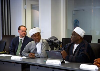 Three men engage in a dialogue at a conference table; two wear traditional attire, the third a suit. One types on a tablet, with nameplates visible—highlighting an International Religious Freedom discussion in Washington D.C.