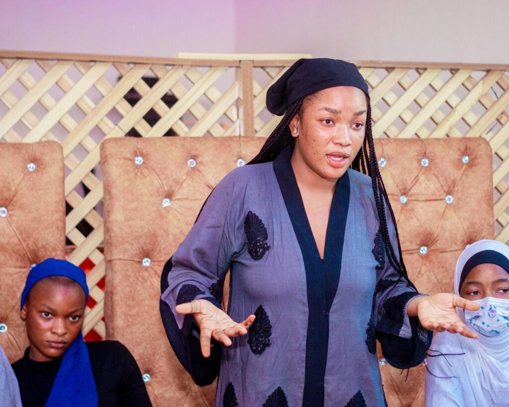 A woman in a dark robe gestures while speaking in Nigeria, standing between two seated women—one in a blue headscarf and the other in a mask and light blue headscarf—as they work to empower communities and protect women.
