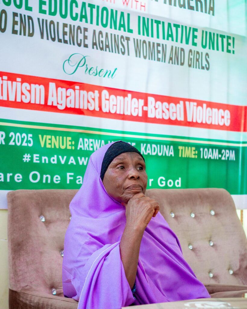 An older woman in a lavender hijab sits in front of a banner at an event in Nigeria, promoting activism to empower communities and protect women and girls from gender-based violence.