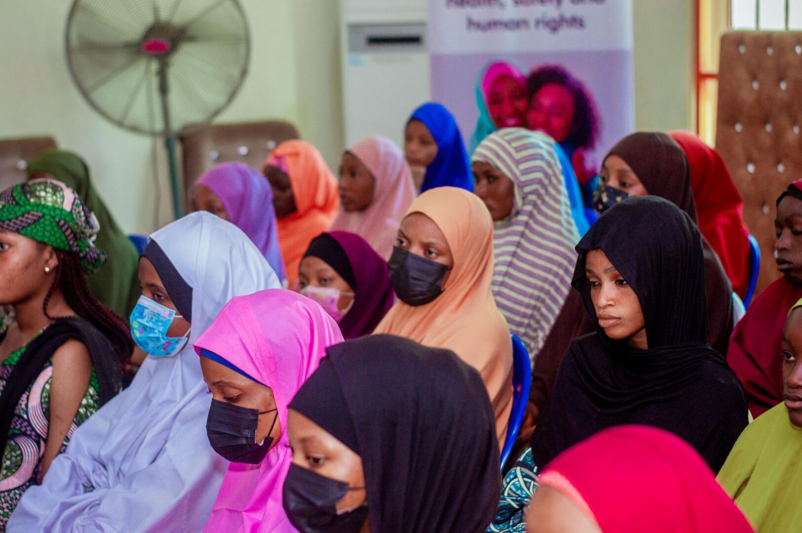 A group of women in Nigeria, some wearing face masks and colorful hijabs, sit attentively in a room during an event focused on empowering communities and supporting women's rights.