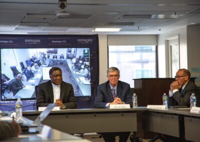 Three men sit at a conference table with nameplates and water bottles in Washington D.C., while a large screen behind them shows a remote video meeting focused on Dialogue and International Religious Freedom.