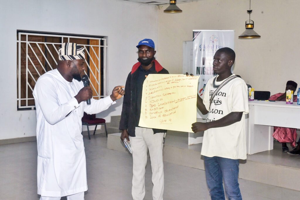 Three men stand indoors in Niger State; one speaks into a microphone, another holds a yellow paper with handwritten notes, and the third stands between them holding a notebook during the International Day of Coexistence.