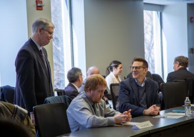 Several people sit and engage in dialogue around a conference table in a brightly lit room with large windows, some discussing topics related to International Religious Freedom, others reviewing documents or devices.