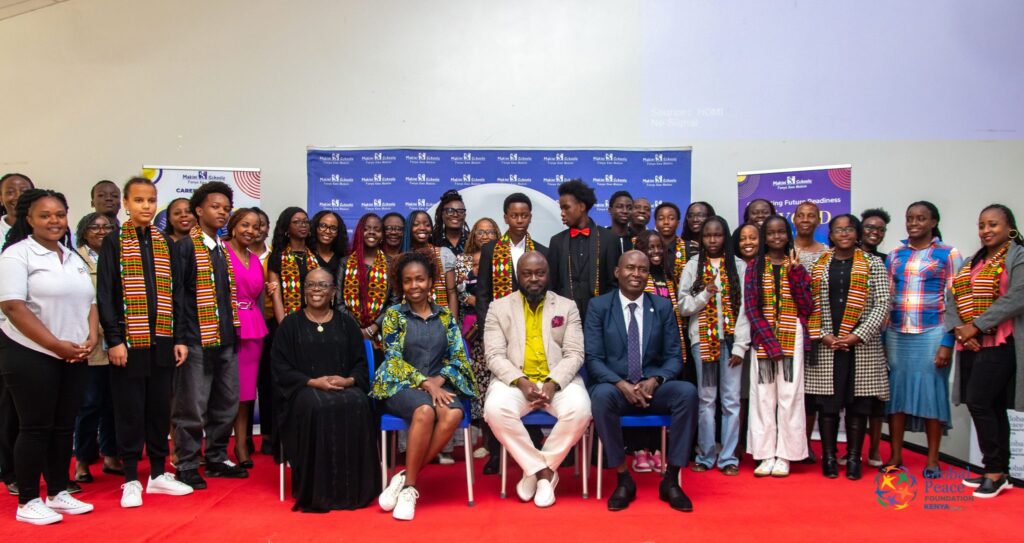 A large group of students and adults pose for a photo on a red carpeted stage, representing youth empowerment through LEAP HUBs. Some are seated at the front and others stand behind them, with GPF Kenya banners visible in the background.