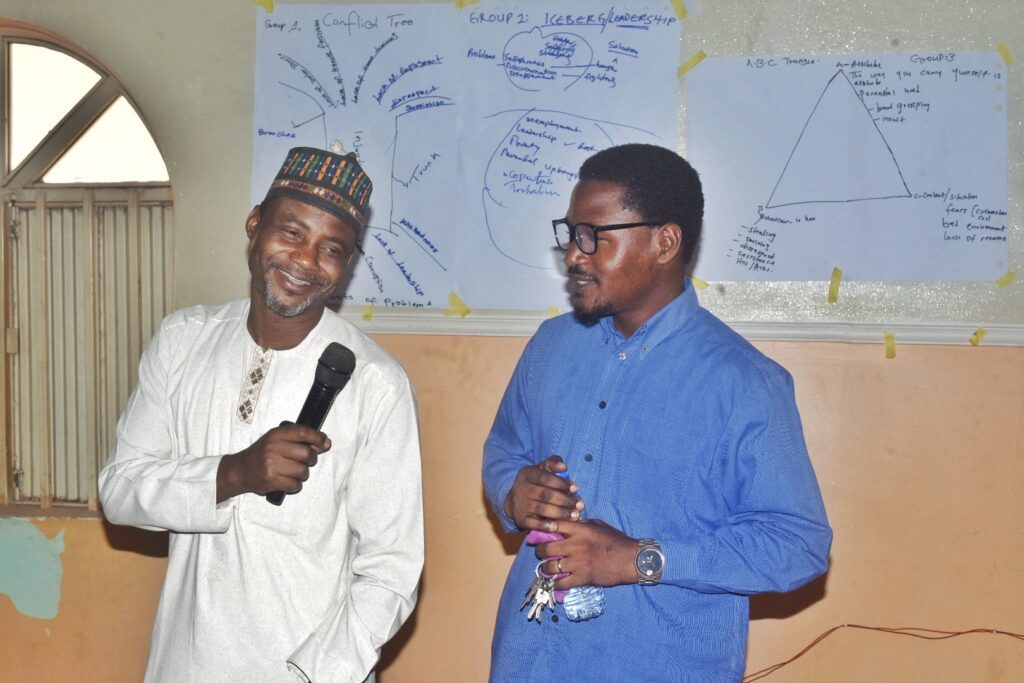 Two men stand indoors; one holds a microphone while speaking about mediation, and the other holds a water bottle. Diagrams and notes related to conflict analysis are posted on the wall behind them.