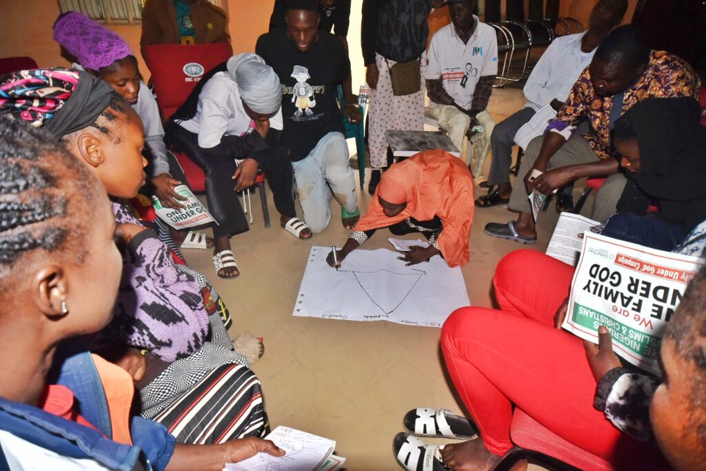 A group of people sit in a circle indoors, watching as one person draws on a large sheet of paper placed on the floor—an interactive session focused on conflict analysis. Some participants hold booklets in their hands.