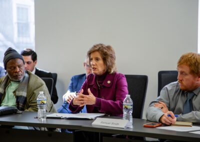 A woman speaks during a Dialogue on International Religious Freedom while seated at a conference table between two men, with water bottles, papers, and nameplates visible in front of them in Washington D.C.