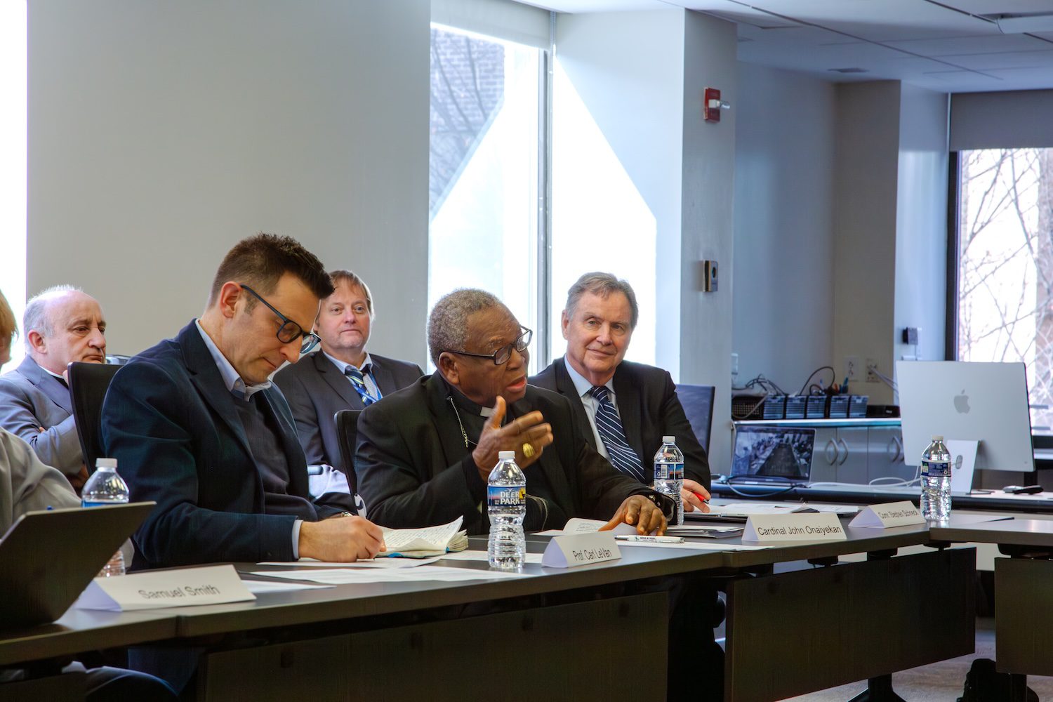 Several people, including Nigerian Faith Leaders, sit at a conference table in a meeting room; one person speaks while others listen and take notes. Bottled water and nameplates are visible on the table as they discuss issues of violence and insecurity.
