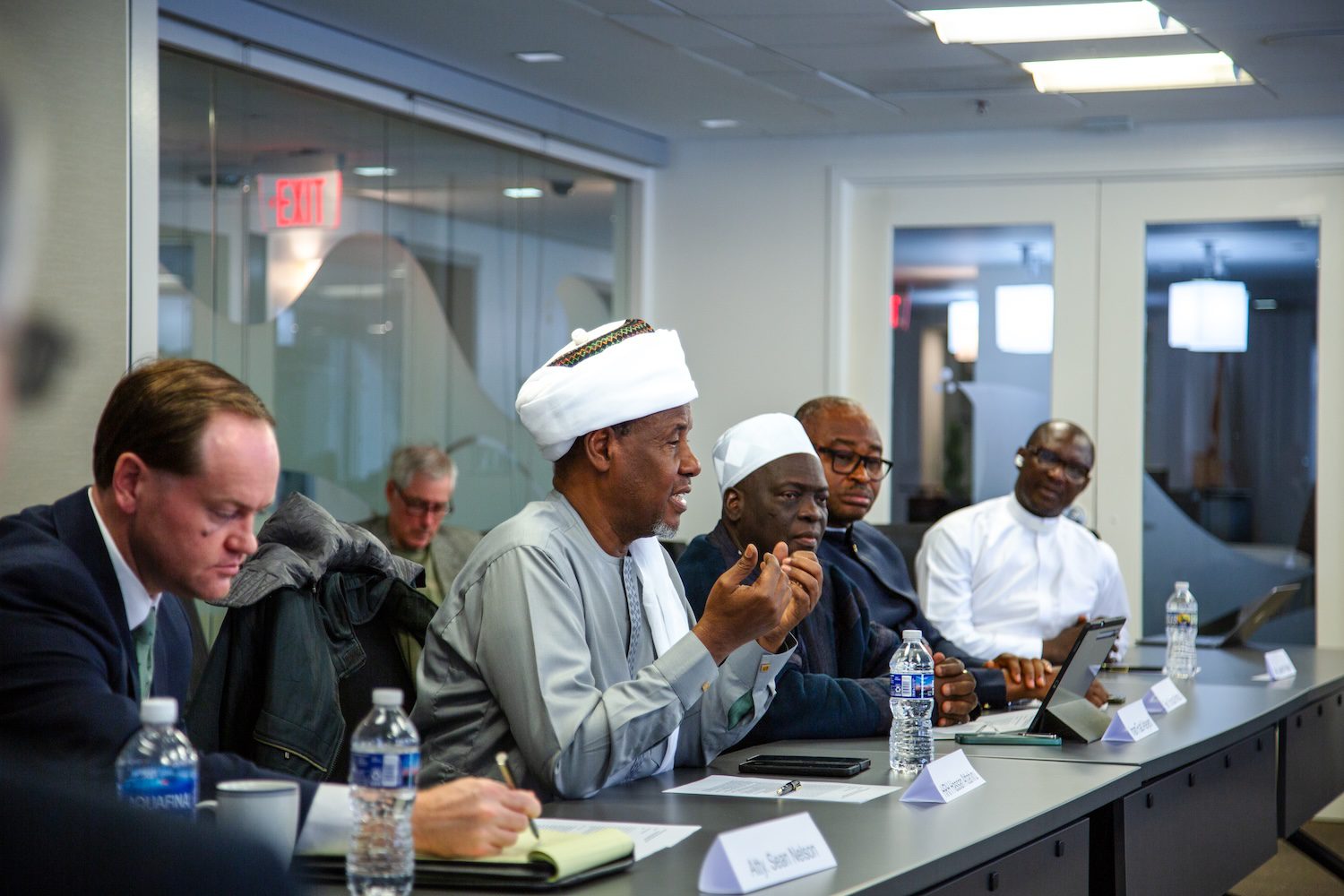 Several men, including Nigerian Faith Leaders, sit at a conference table during a meeting. One man in traditional attire speaks on insecurity and violence while others listen; laptops, water bottles, and papers are visible on the table.