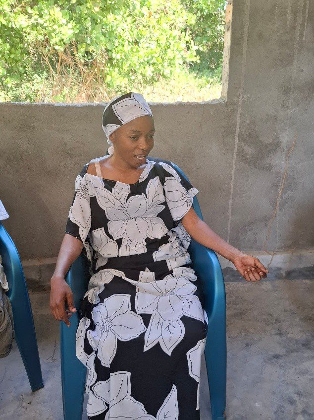 A woman in a black and white floral dress and headscarf sits in a blue plastic chair indoors, with a window and greenery behind her—reflecting GPF Tanzania’s commitment to grassroots peacebuilding within communities.