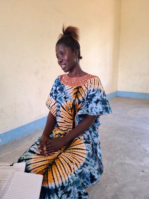 A woman in a brightly patterned dress sits on a chair in a sparsely furnished room, with papers and a notebook visible in the foreground—reflecting her commitment to grassroots peacebuilding.