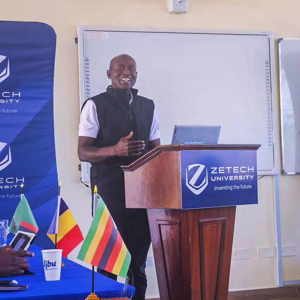 A man stands at a podium giving a presentation to youth at Zetech University. Several international flags and a laptop are on the podium, with a whiteboard in the background, celebrating future leaders and peacebuilders.