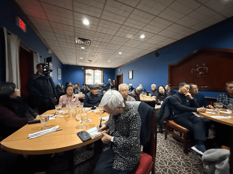 A group of adults sits around tables in a restaurant or meeting room, listening attentively while some take notes; a camera crew is present in the background, possibly discussing external information about North Korea.