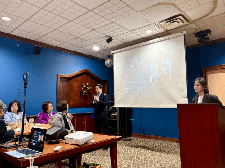 A man speaks at a microphone in a meeting room with attendees seated around tables; a woman stands at a podium, and a projector displays a presentation slide about North Koreans and the impact of external information.