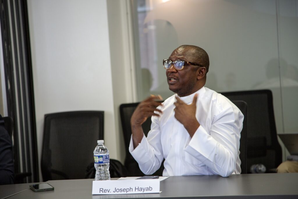 A man in glasses and a white shirt, identified by a nameplate as Rev. Joseph Hayab, speaks at a conference table about International Religious Freedom, with a water bottle and phone in front of him.