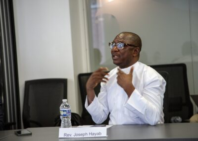 A man in glasses and a white shirt, identified by a nameplate as Rev. Joseph Hayab, speaks at a conference table about International Religious Freedom, with a water bottle and phone in front of him.