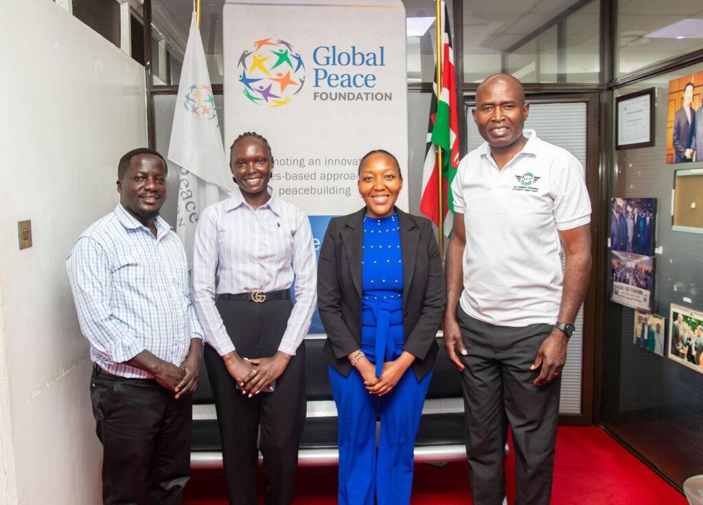 Four people stand indoors in front of a Global Peace Foundation banner and flag, posing for a group photo that highlights their commitment as youth leaders dedicated to peacebuilding.