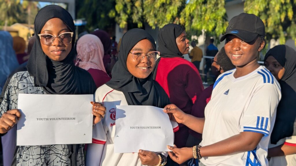 Three young women wearing hijabs stand outdoors in sunlight, holding papers that read