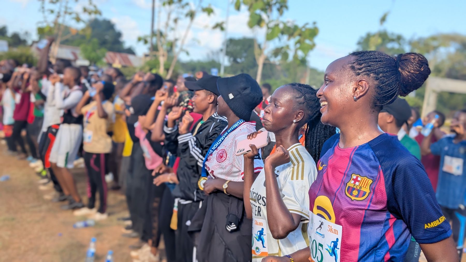 A group of runners, including participants in women’s leadership and youth volunteerism initiatives, gathers outdoors before a race, some smiling and wearing athletic gear and numbered bibs; trees and blue sky are visible in the background.