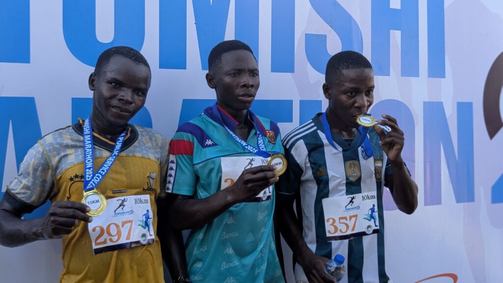 Three men wearing running bibs pose with medals at a marathon event, promoting youth volunteerism as they proudly display their achievements in front of an event backdrop.