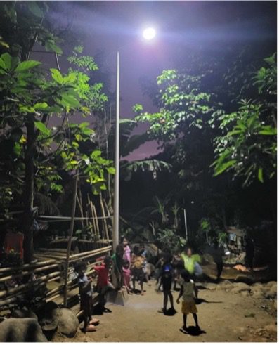A group of children stand and play under a bright streetlight at night, surrounded by trees and vegetation in a rural All-Lights Village—a vibrant scene reflecting community development supported by GPF Philippines.