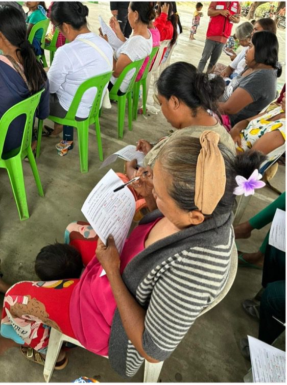 A group of women seated on plastic chairs fill out forms during a community development meeting in an outdoor covered area, part of the GPF Philippines' All-Lights Village initiative.