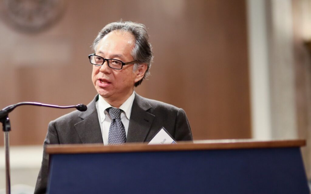 A man in a suit and tie speaks at a podium with a microphone in a formal indoor setting.
