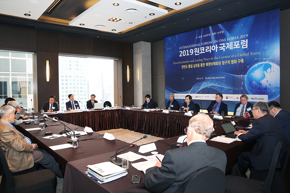 A group of people in formal attire are seated around a large conference table during the 2019 International Forum on One Korea in a modern meeting room.