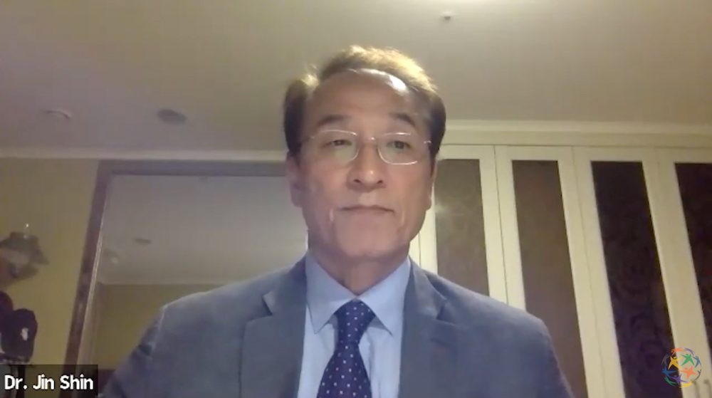 A man in a suit and tie speaks during a video call. The overlay text reads 