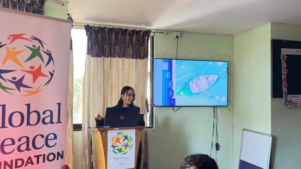 A woman stands at a podium giving a presentation, with a slide showing a diagram on a screen, next to a Global Peace Foundation banner at the Global Peacebuilders Leadership Program, inspiring young leaders from GPF Nepal.