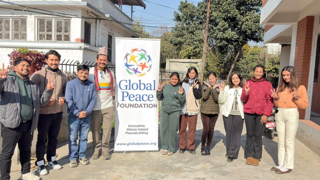 A group of people stand outdoors beside a Global Peace Foundation (GPF Nepal) banner, smiling and making peace signs with their hands, celebrating the spirit of Young Leaders from the Peacebuilders Leadership Program.