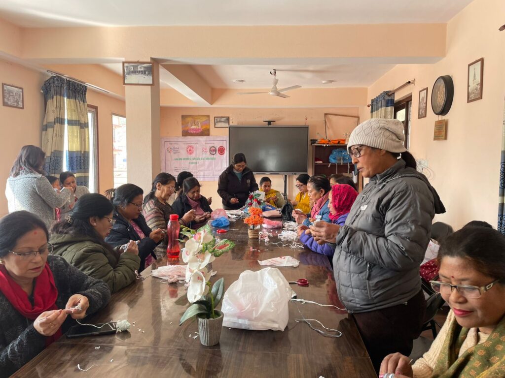 A group of women sit and stand around a table, knitting and working with yarn in a bright room decorated with posters and plants, reflecting GPF Nepal’s commitment to women empowerment and a greener Kathmandu.