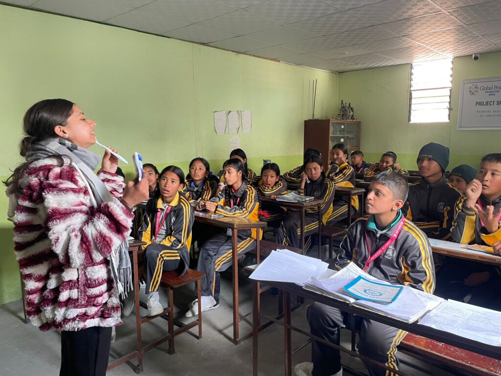 A teacher stands at the front of a classroom, holding a marker and speaking to students in uniform who are seated at desks, attentively listening—a scene reflecting GPF Nepal’s commitment to women empowerment through education.