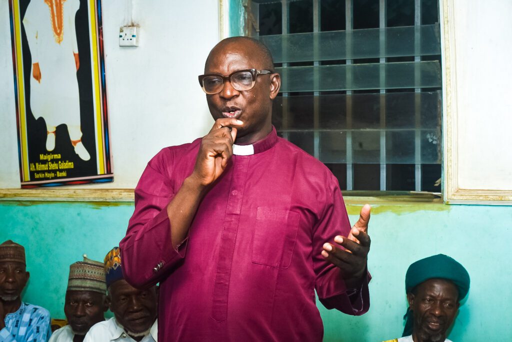 A man in a maroon clerical shirt speaks while gesturing with his hand during a Community-Led Peace event; several people sit and listen in the background at GPF Nigeria.