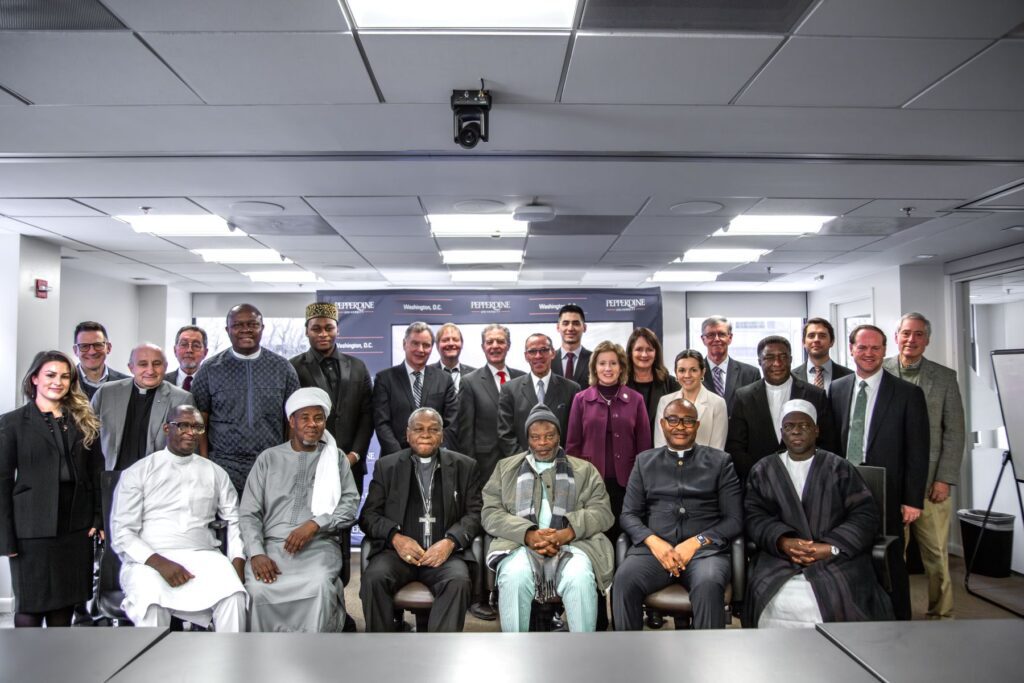 A group of men and women, including interfaith leaders, pose for a formal photo in a conference room, with a banner reading