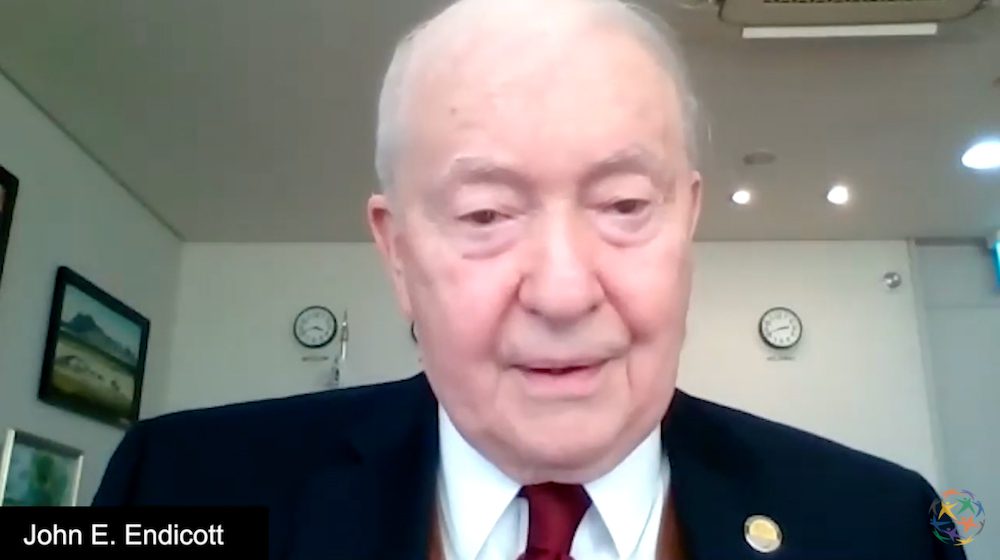 An older man in a suit and tie speaks on a video call. The name 