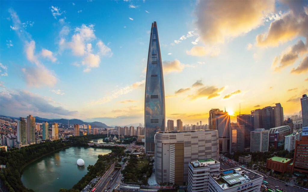 Tall skyscraper surrounded by modern buildings and a lake at sunset, with partly cloudy sky and cityscape in the background.