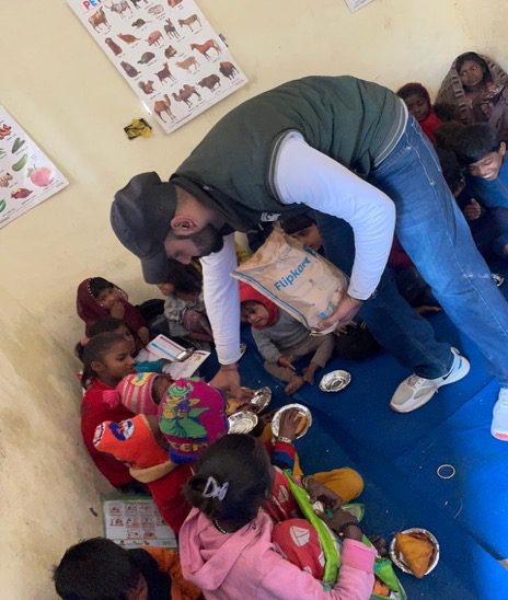A man distributes food to a group of children seated on the floor in a classroom adorned with educational posters, highlighting community engagement and the spirit of Republic Day 2026.
