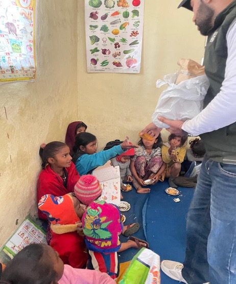 A group of children sit on the floor near a wall, receiving food and supplies from an adult in a room decorated with educational posters, highlighting community engagement as part of Republic Day 2026 celebrations.