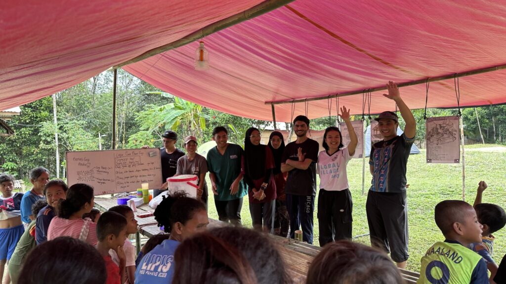 A group of adults stand under a red canopy in Kuala Lipisment, some waving, while children sit or stand facing them. Whiteboards with writing are visible in the background—an outdoor week of grit and giving amid lush greenery.