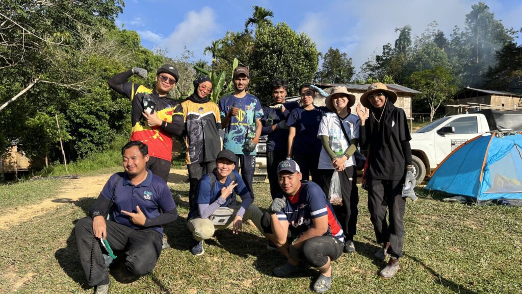 A group of people poses outdoors on grass near a tent, a white pickup truck, and trees at Ulu Semul, with some giving peace signs and smiling at the camera.