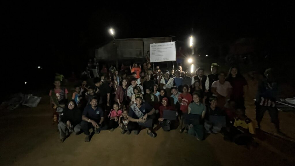 A large group of people, including children and adults, pose for a photo at night on the dirt ground in front of a lit sign and building during the week of giving in Ulu Semul, Kuala Lipisment.