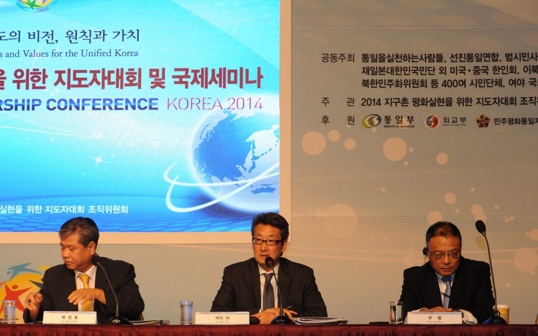 Three men in suits are seated at a conference table with microphones in front of a backdrop displaying the text