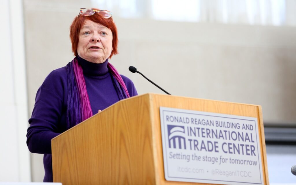 An older woman with short red hair speaks at a podium labeled