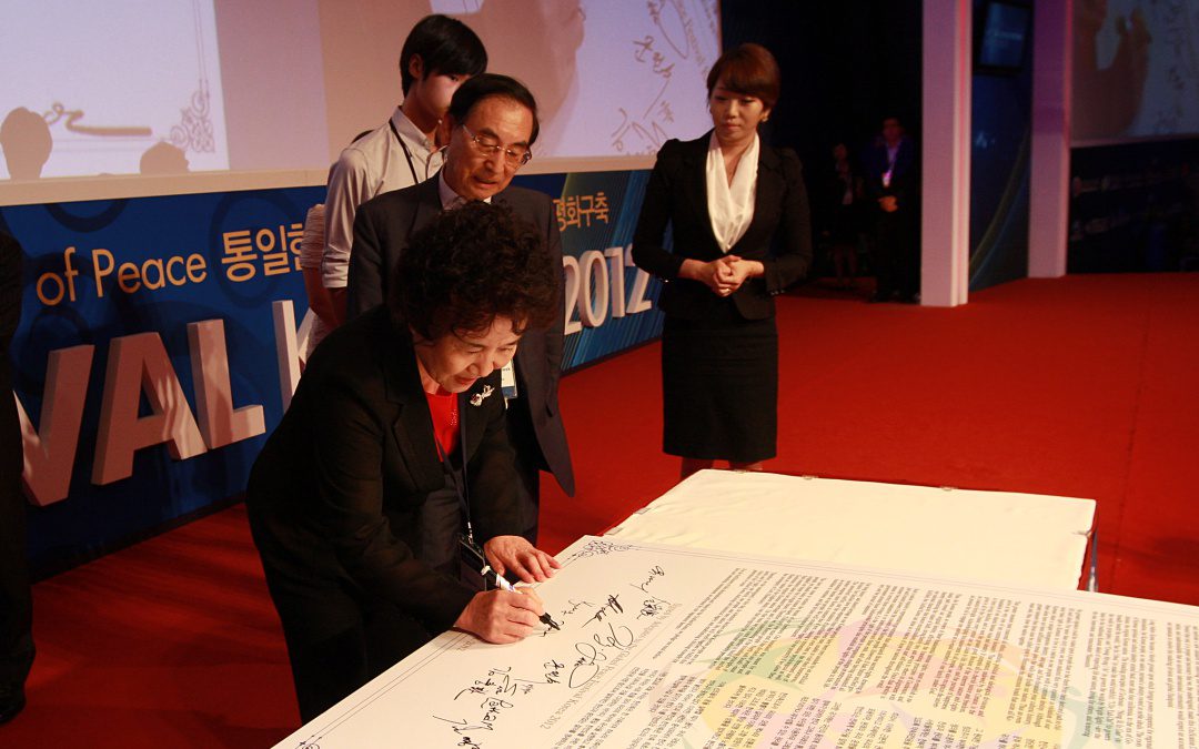 A woman signs a large document on a table while three people observe at an indoor event with a red carpet and blue banners.