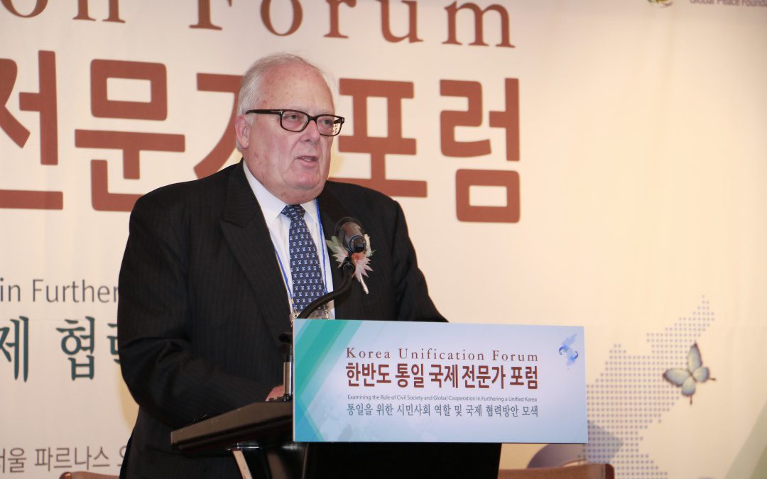 An older man in a suit and tie speaks at a podium during the Korea Unification Forum, with a sign in Korean and English displayed on the podium.
